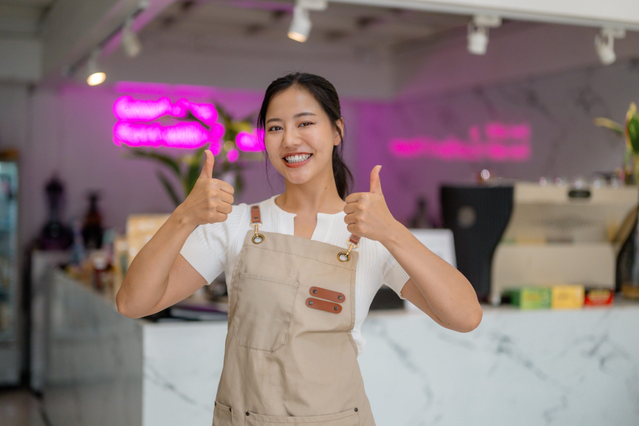 Young Asian female barista giving thumbs up in coffee shop